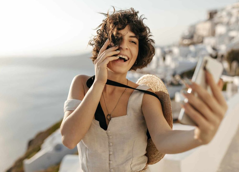 brunette-curly-woman-in-beige-dress-and-straw-hat-smiles-sincerely-and-takes-selfie-outside-in-old.jpg